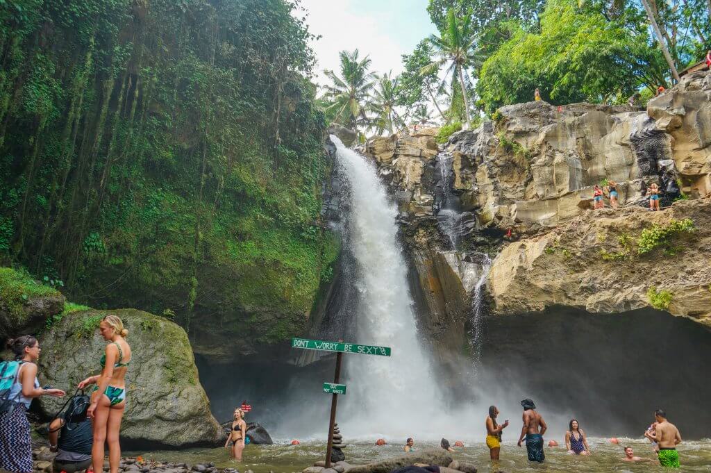 people stand at the foot of a Bali waterfall. A sign in the water says "don't worry, be sexy. But not naked."