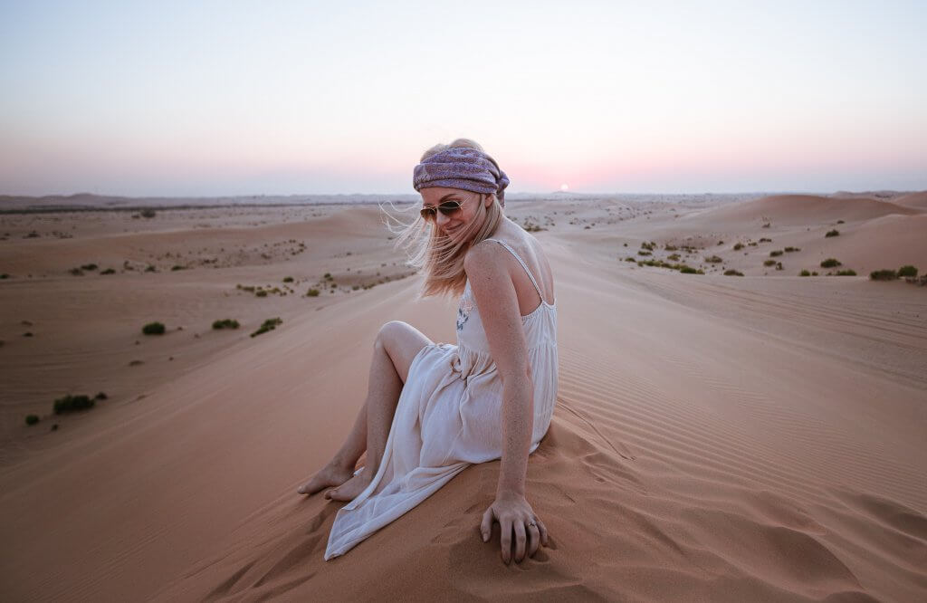 blonde woman sitting atop a sand dune wearing a headscarf, white dress, and sunglasses looking back at the camera